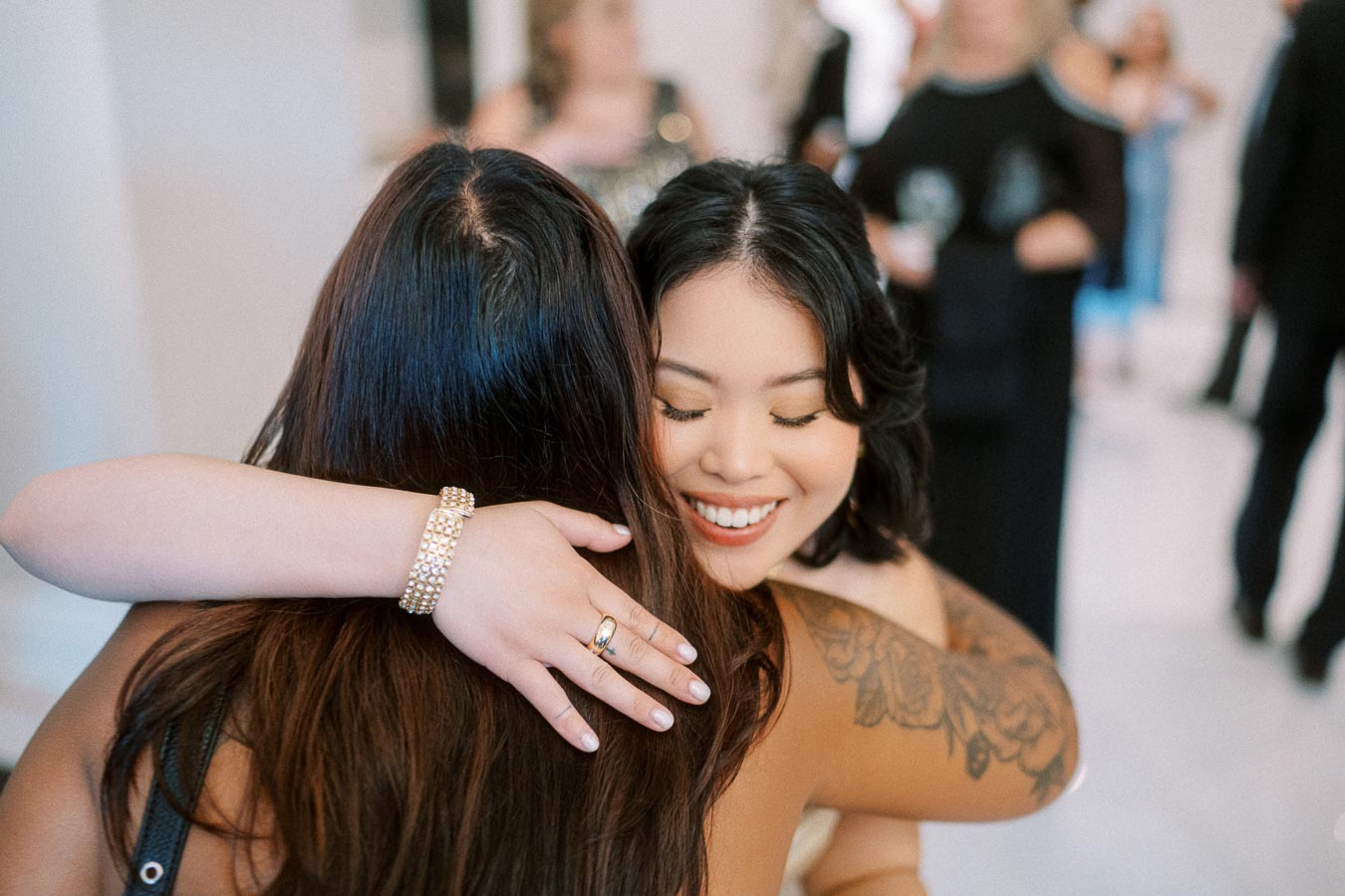 Two women embracing warmly at an elegant event, one with a smile and tattoos, the other adorned with a sparkling bracelet,