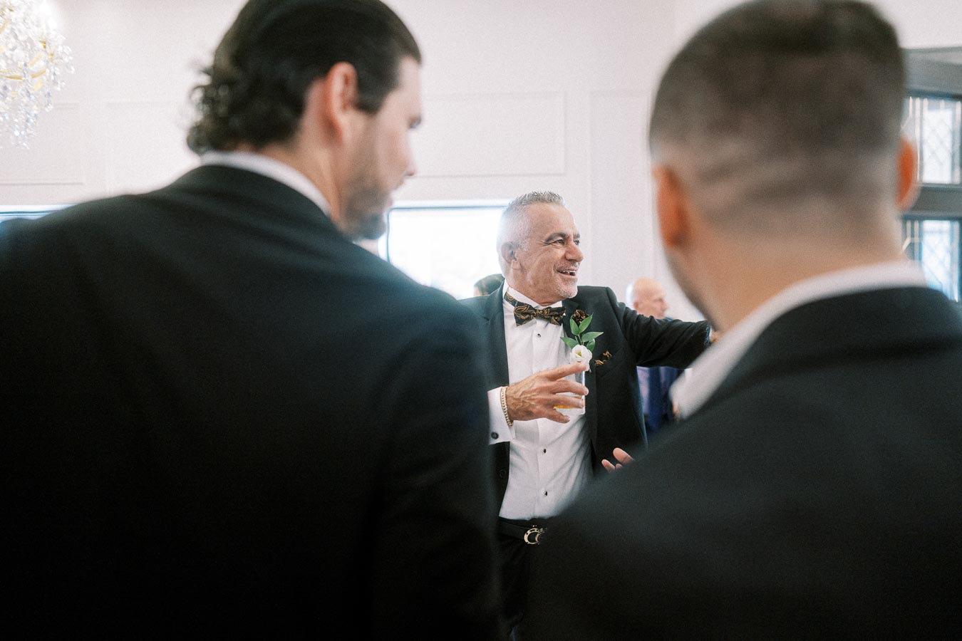 A joyful older man in a tuxedo with a bow tie holding a drink, engaging with two other formally dressed men at an elegant