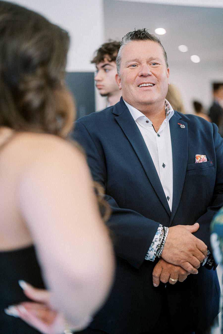 Man in a navy suit smiling and engaged in conversation at a social event, with a blurred woman in the foreground.