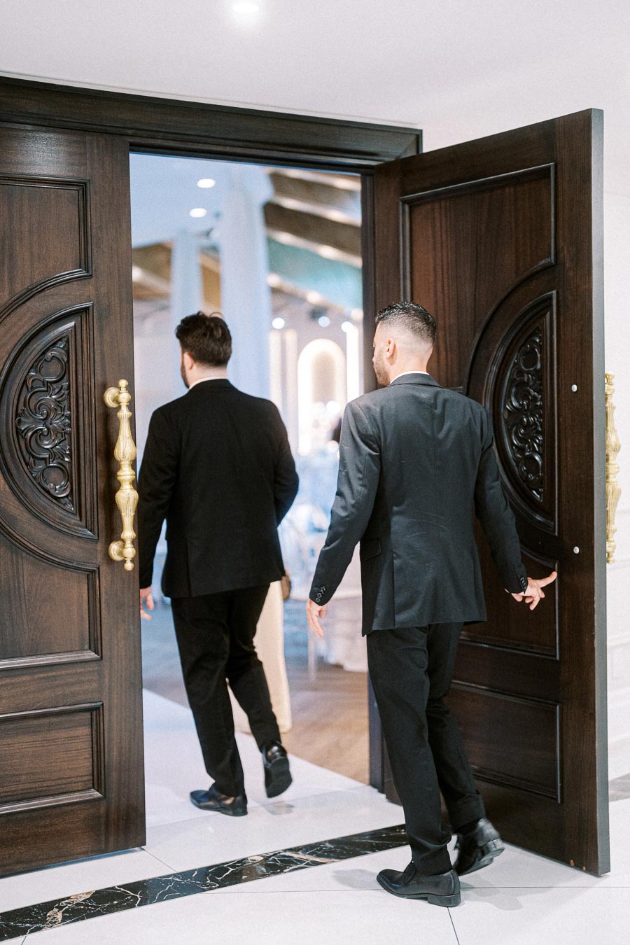Two men in formal black suits walking through a decorative wooden door into a brightly lit room, suggesting an entrance to