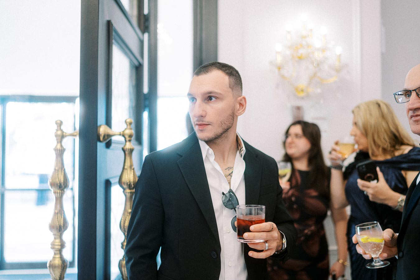 A man in a formal black suit holds a drink at a social gathering, surrounded by other guests who are chatting and holding