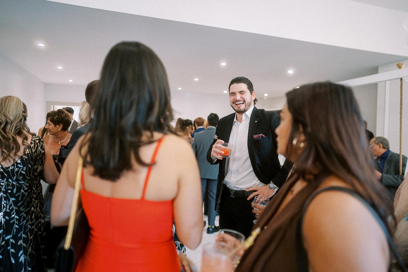 A lively social gathering with people in formal attire, including a man in a suit laughing and holding a drink, creating a