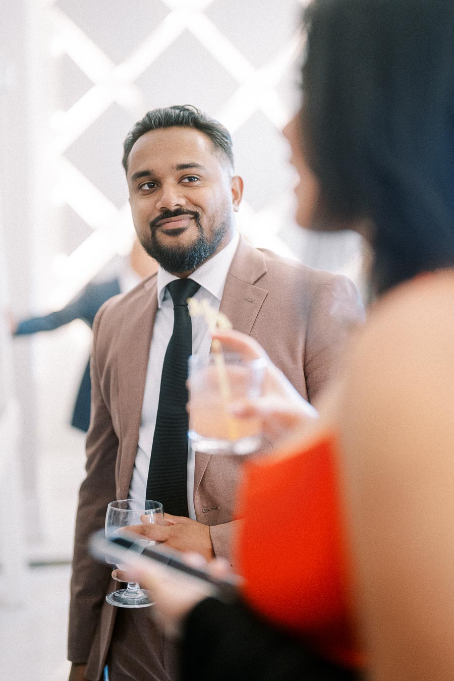 A well-dressed man in a brown suit at a social gathering, holding a glass and smiling, with a blurred background and a