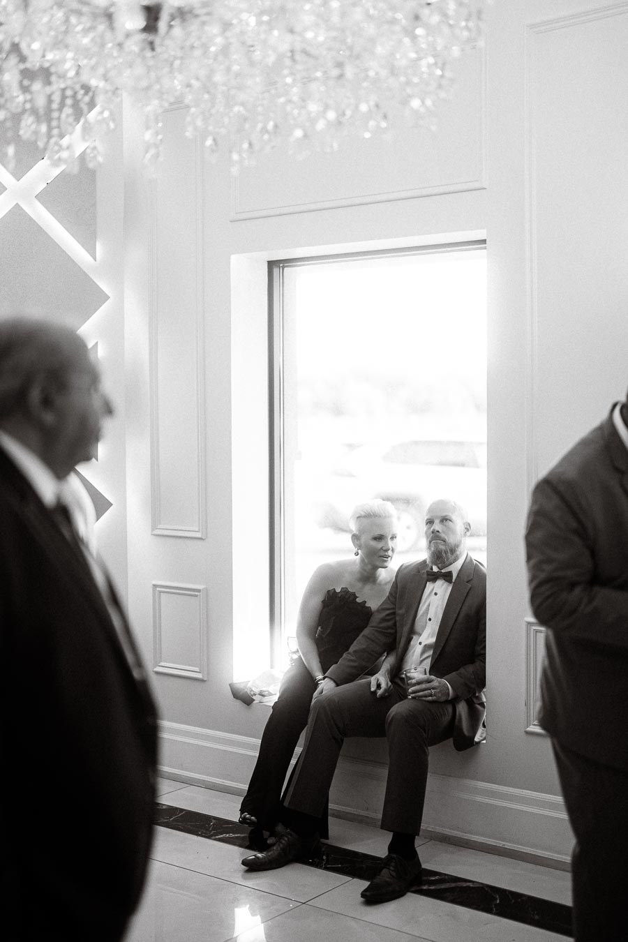 Black and white photo of a couple dressed formally, sitting by a window at an elegant event, surrounded by other guests.