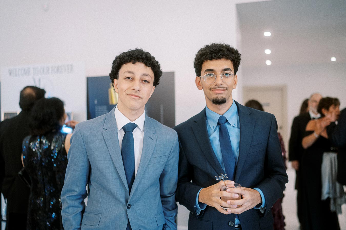 Two young men in suits standing at a formal event setting, with one holding a drink and people socializing in the background.