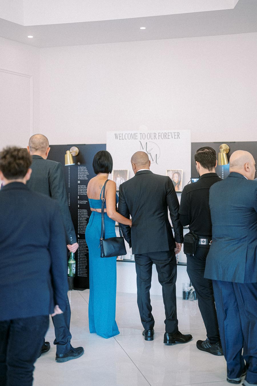 A group of elegantly dressed people stand in front of a wedding welcome sign, reading Welcome to Our Forever, at a stylish
