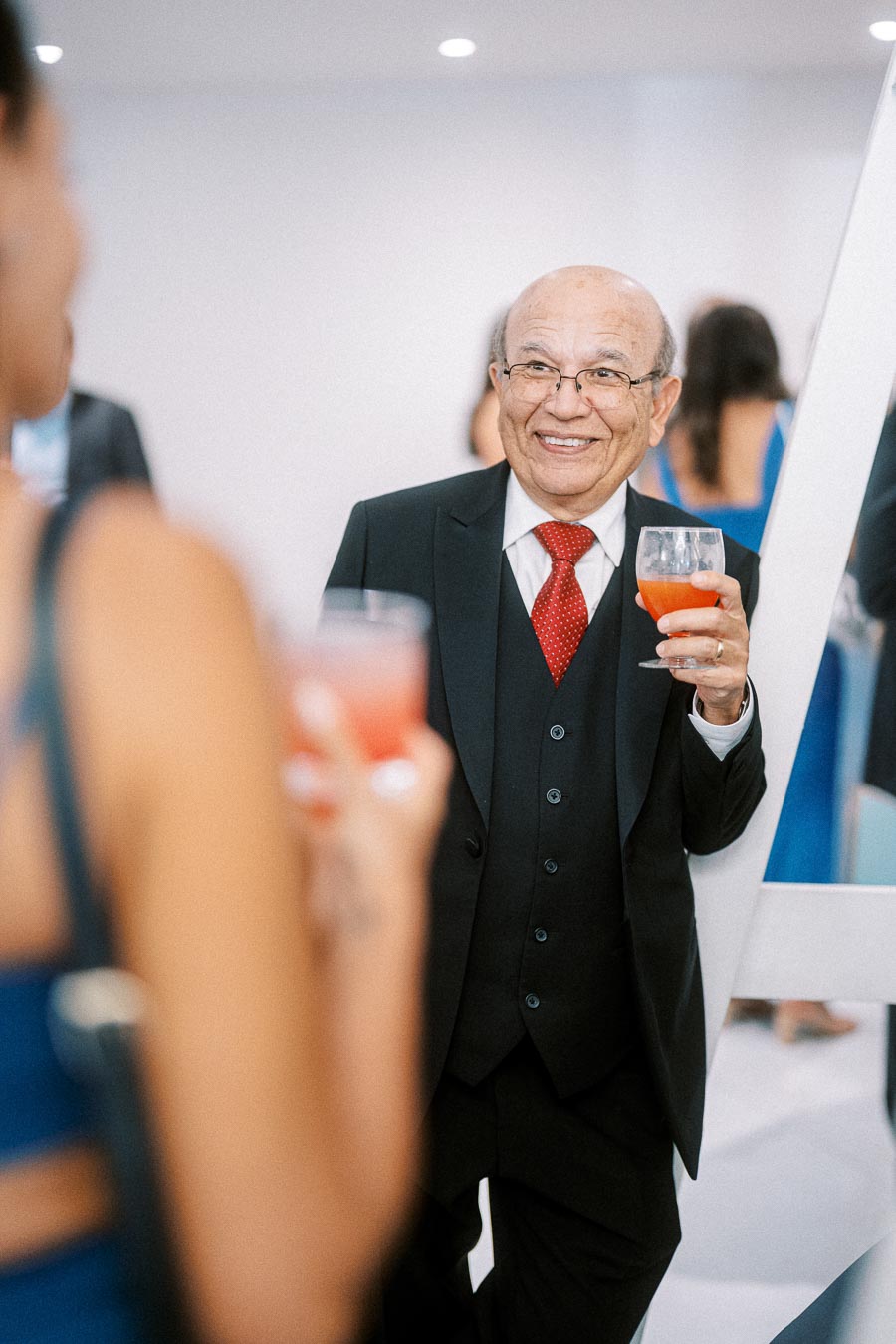 Elderly gentleman in a suit holding a red cocktail drink, smiling at a social event.