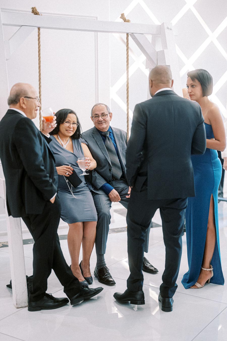 A group of elegantly dressed people socializing at a formal event, with some seated on a swing and others standing, enjoying