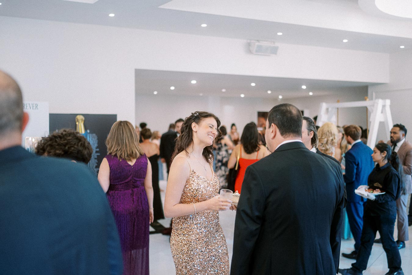A group of elegantly dressed people socializing at a formal event, with a woman in a sparkling gold dress smiling while