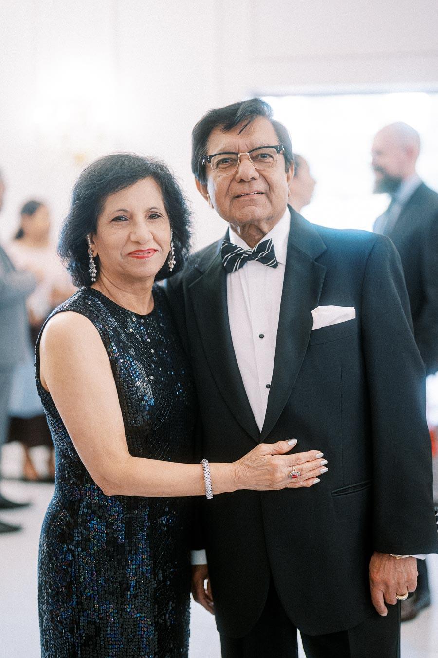 Elegant couple dressed in formal attire, woman in a sequined dress and man in a tuxedo with bow tie, smiling at a