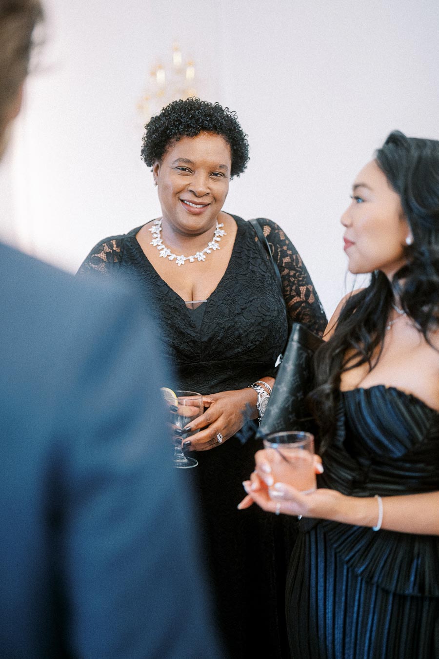 Two women in elegant black dresses holding drinks at a formal event, engaging in conversation and smiling.