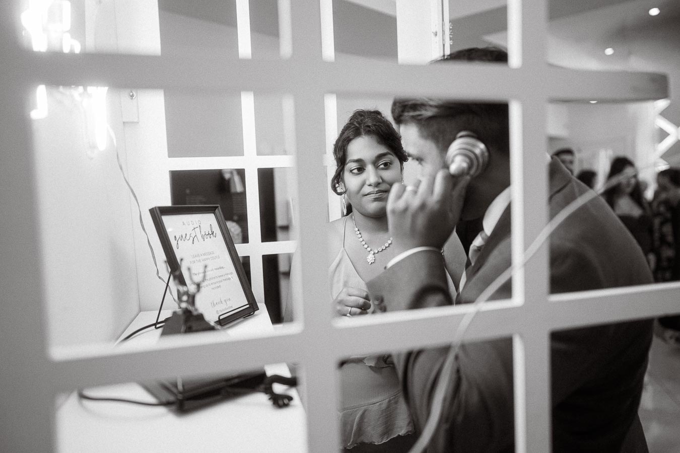 A man in a suit listens to an audio guest book message while a woman looks on, captured in a black and white photo through a
