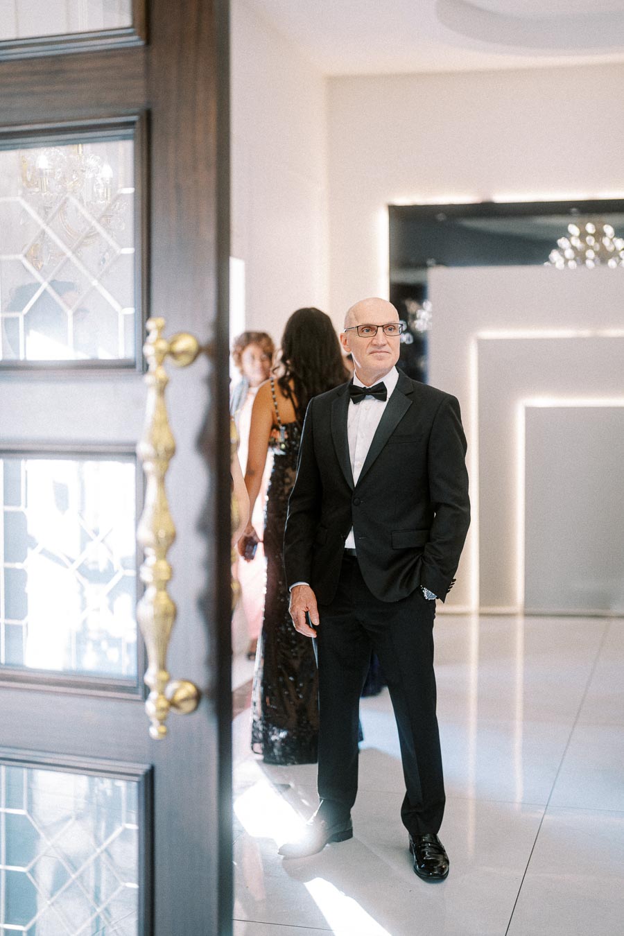 Elegant gentleman in a black tuxedo at a formal event entrance, with blurred attendees in the background and lavish interior