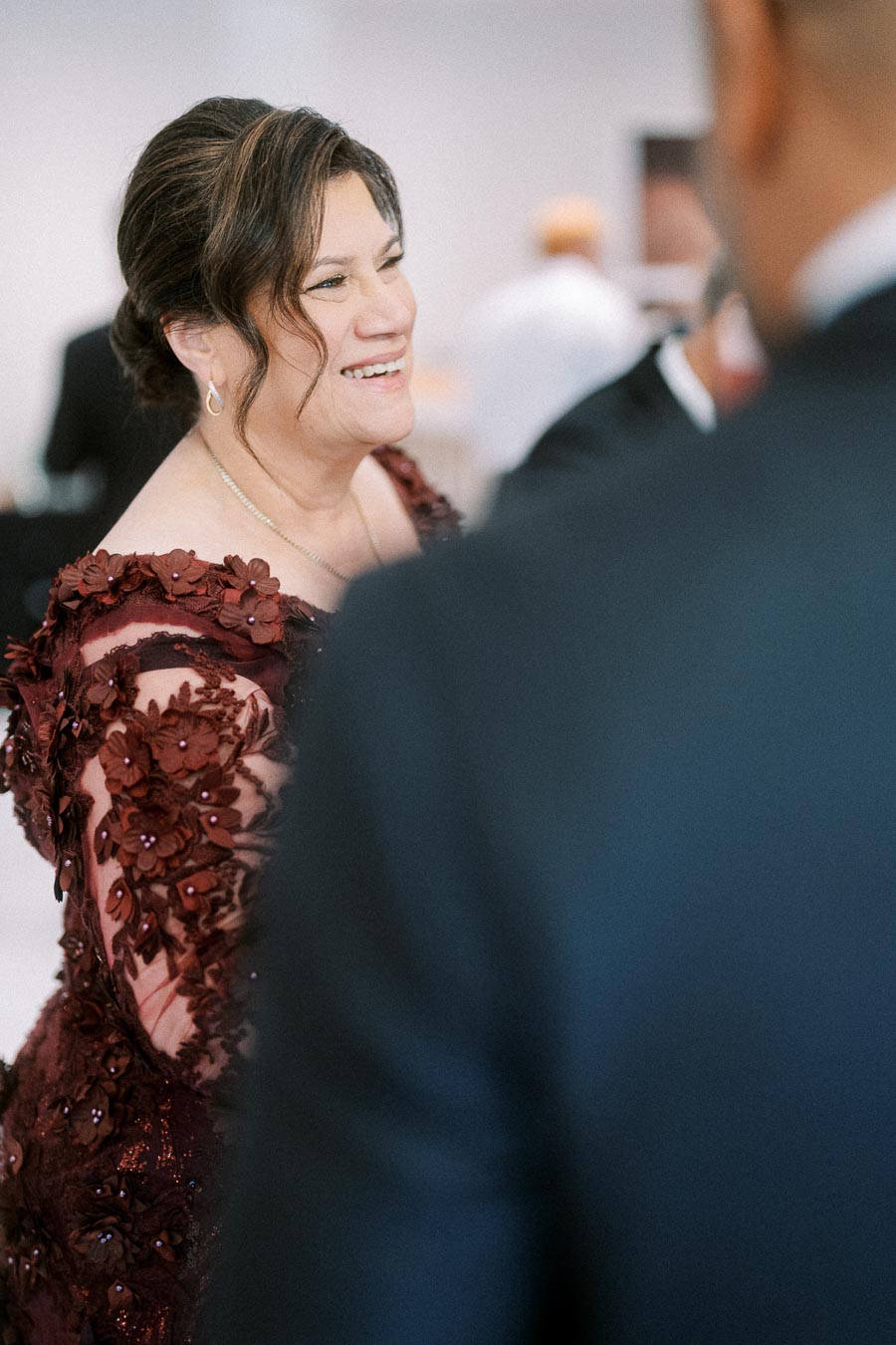 A woman in a burgundy floral lace dress smiling at an event with people in the background.