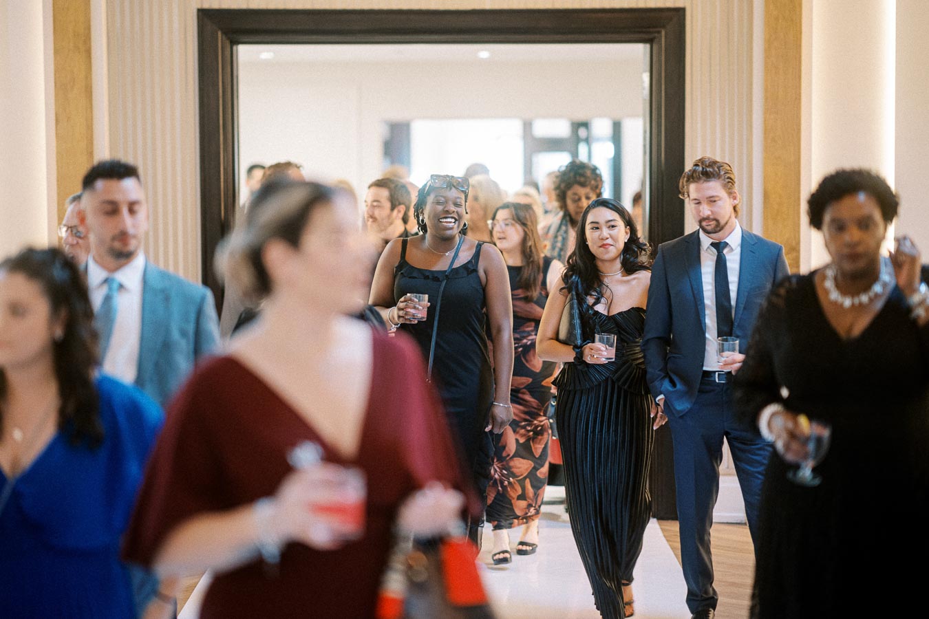 A diverse group of well-dressed individuals socializing at a formal event, walking through a stylish hallway while holding