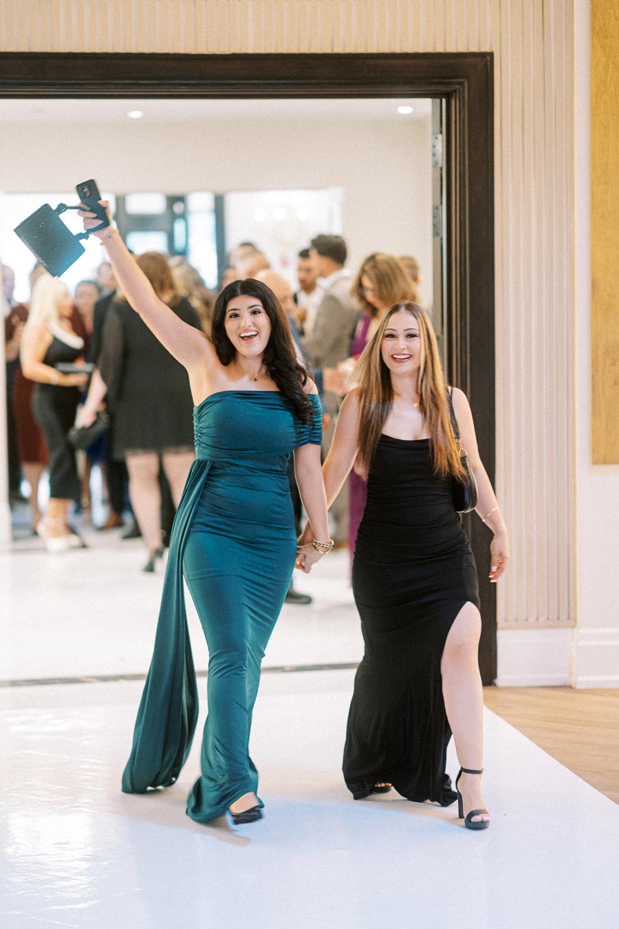 Two women in elegant evening gowns, one teal and one black, walk hand-in-hand at a formal event, smiling and celebrating.