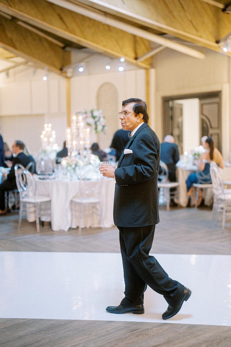 A man in a suit holding a drink at an elegantly decorated event venue, with tables adorned with flowers and candelabras in
