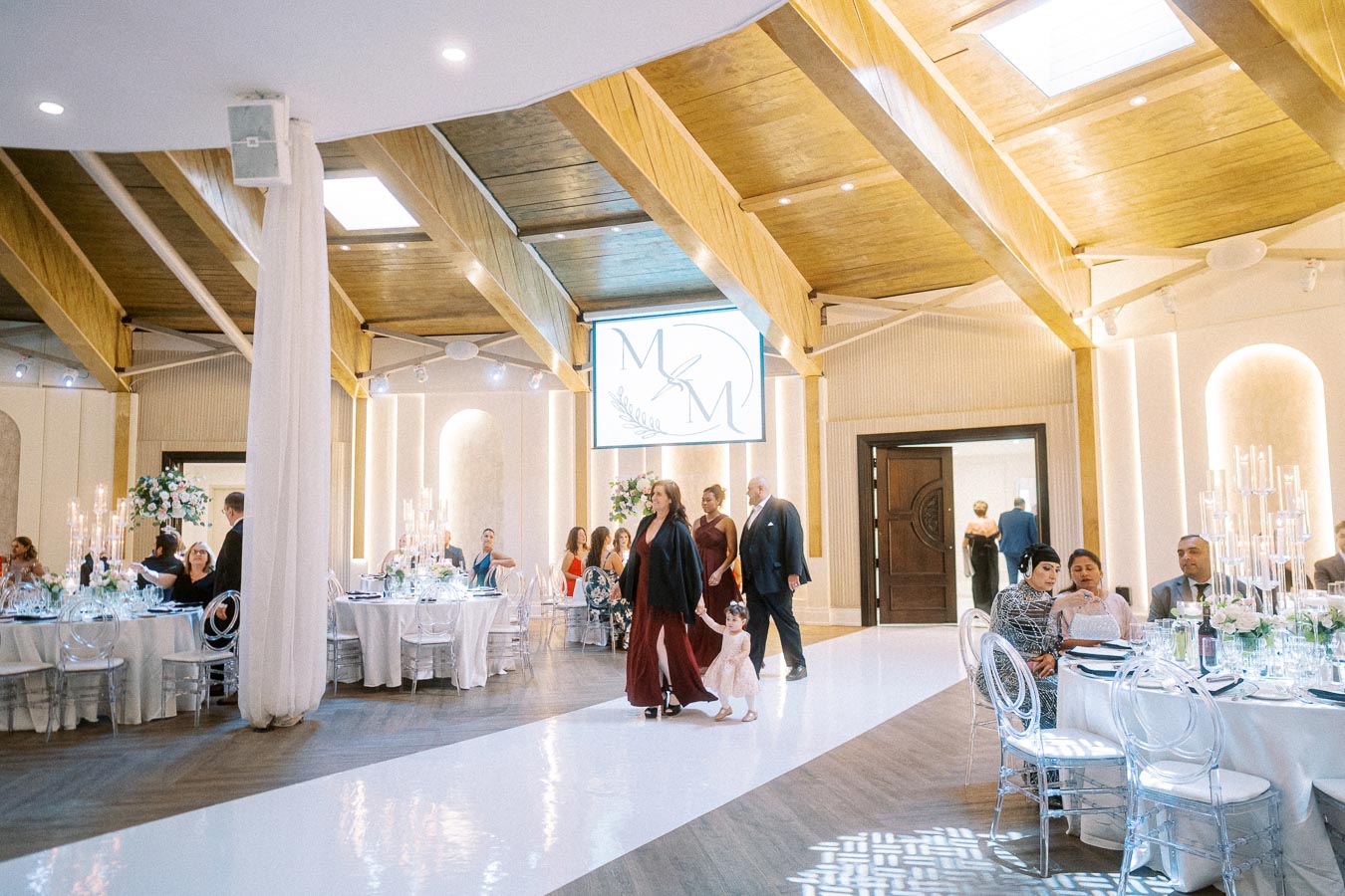 Elegant wedding reception venue with wooden ceilings and modern decor. Guests seated around round tables adorned with white