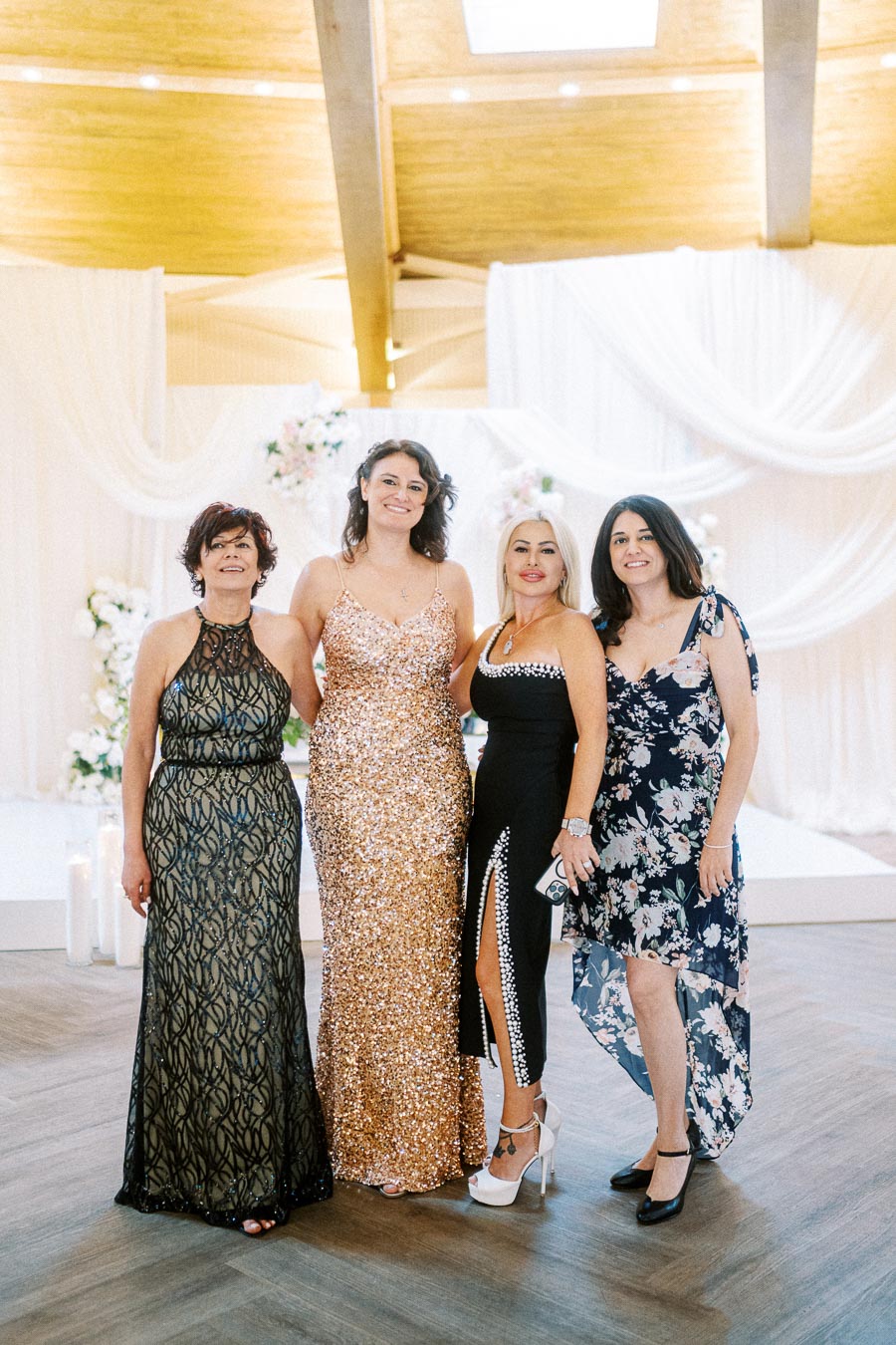 Four women elegantly dressed, posing together at a formal event surrounded by soft drapery and flowers.