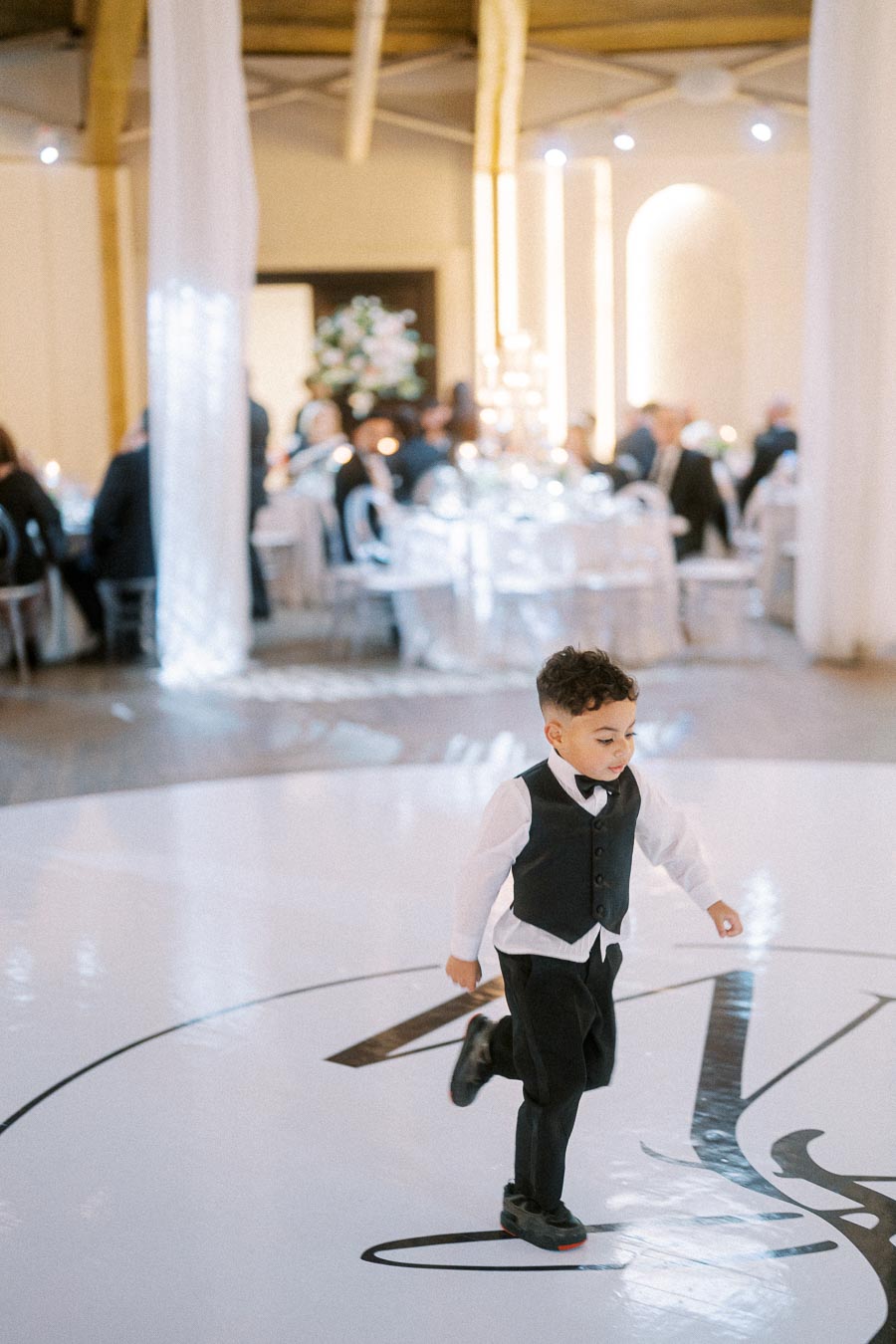 A young boy in a formal black vest and bow tie runs across a glossy dance floor at a wedding reception, with elegantly set