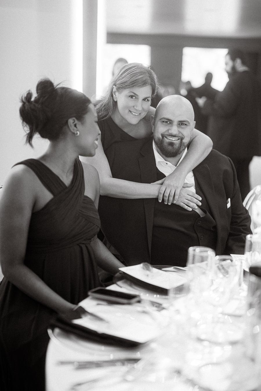 A joyful group at an elegant event, with a woman standing and embracing two colleagues seated at a table set for dinner. The