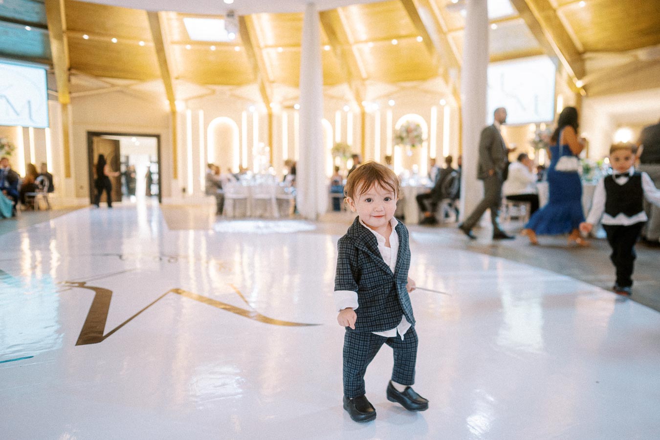A young child in a stylish suit walks confidently on a polished ballroom floor during a lively wedding reception, surrounded