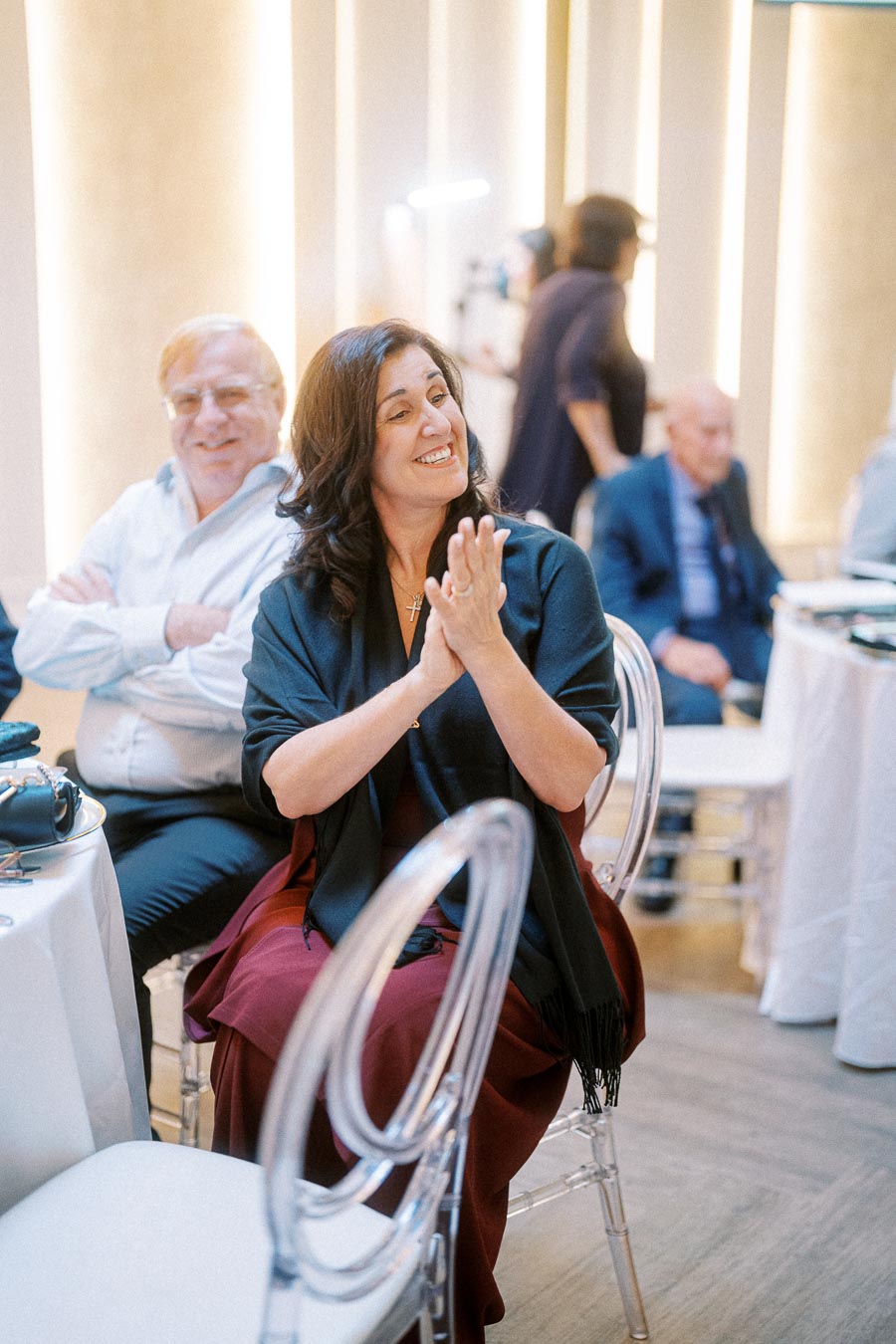 Happy woman clapping at a formal event, seated beside a smiling man; both appear engaged and enjoying the occasion.