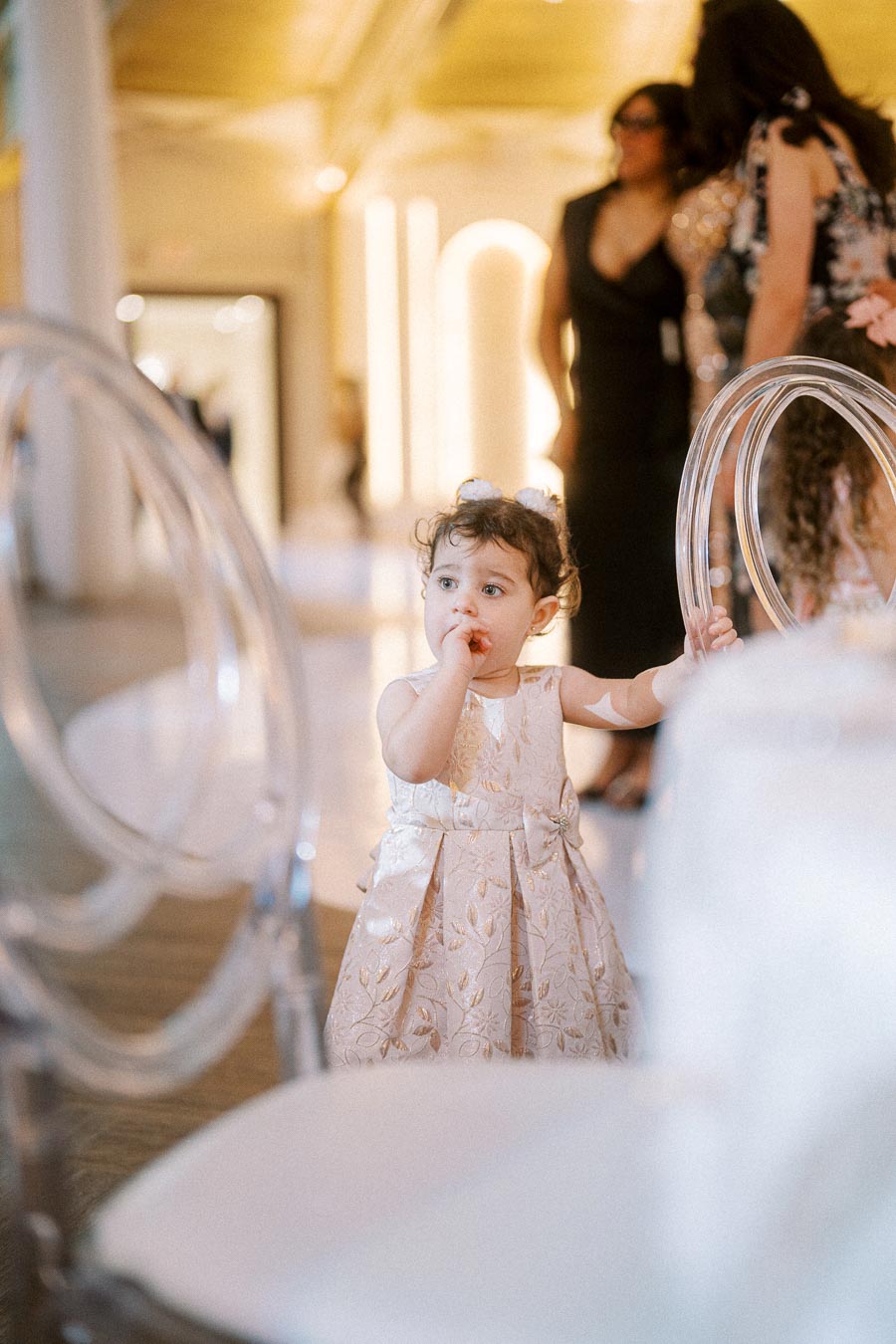 A toddler in an elegant floral dress stands near clear chairs at an indoor event, with soft lighting and blurred figures in