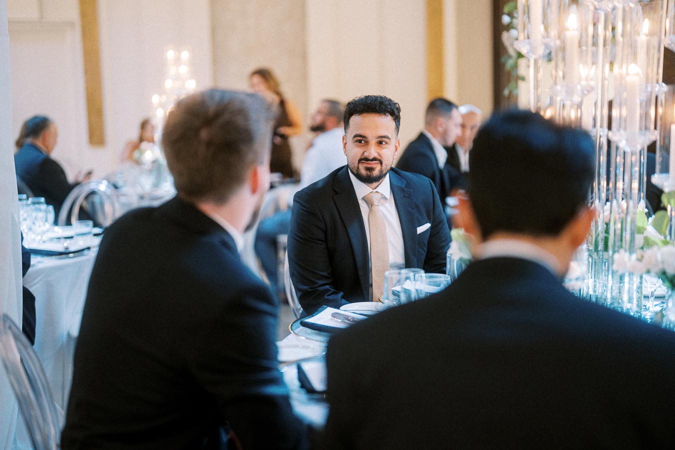 A man in a suit sits at an elegantly decorated table during a formal event, conversing with others in a sophisticated