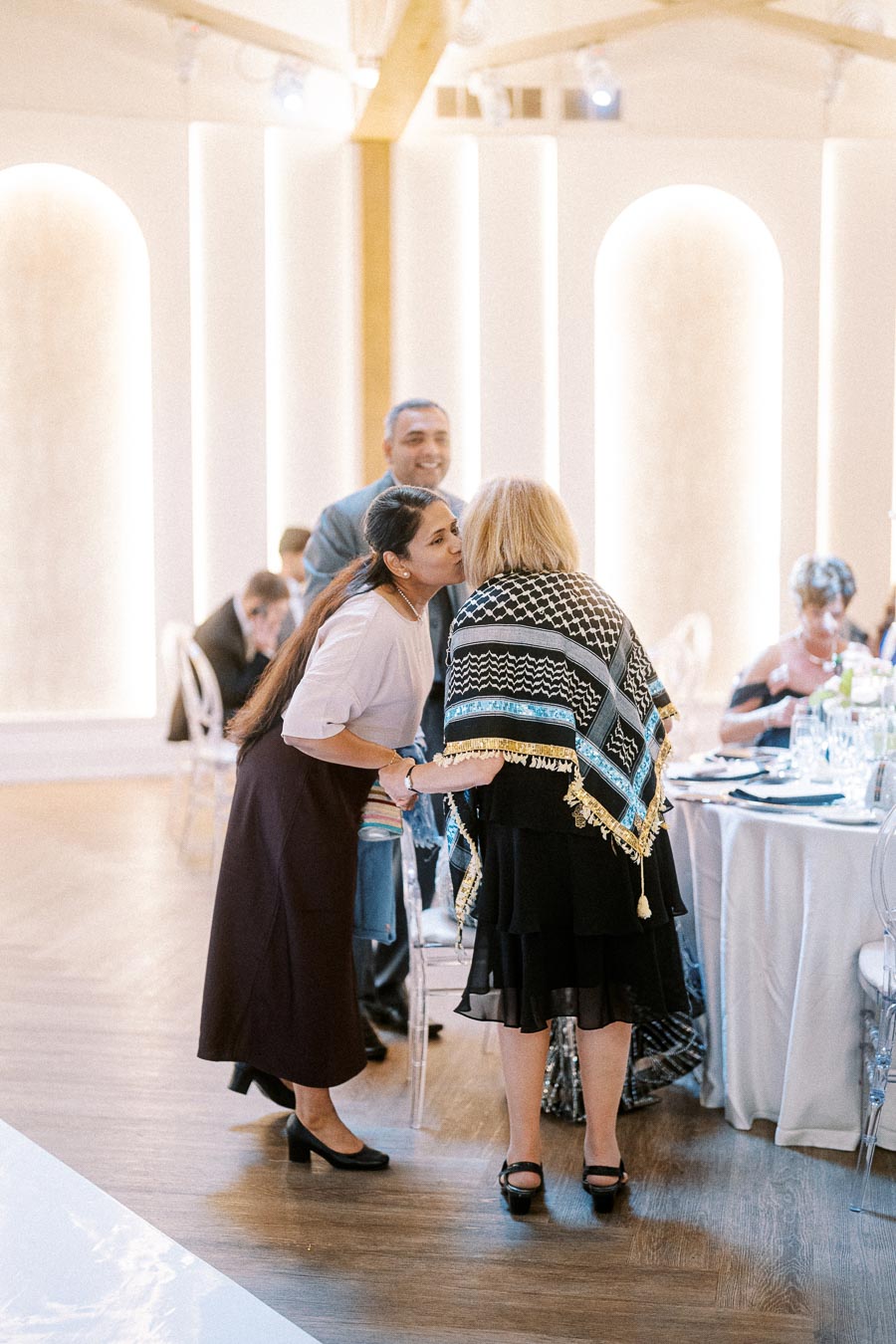 A woman greeting an older woman with a kiss on the cheek at a formal event, with tables elegantly set in the background,
