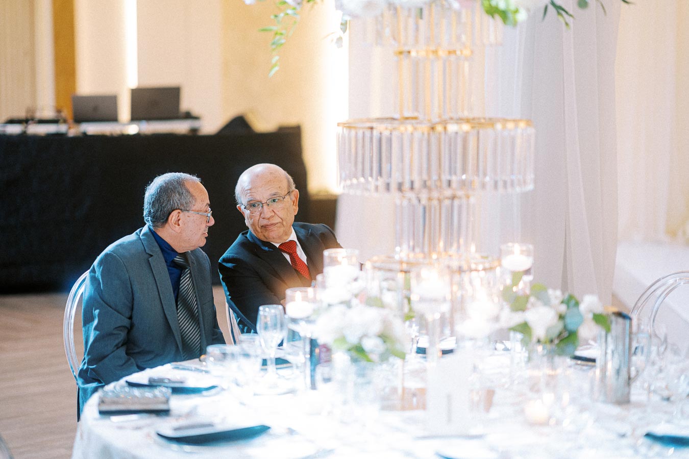 Two elderly men in formal attire sit together at an elegantly decorated banquet table, conversing under soft lighting, as