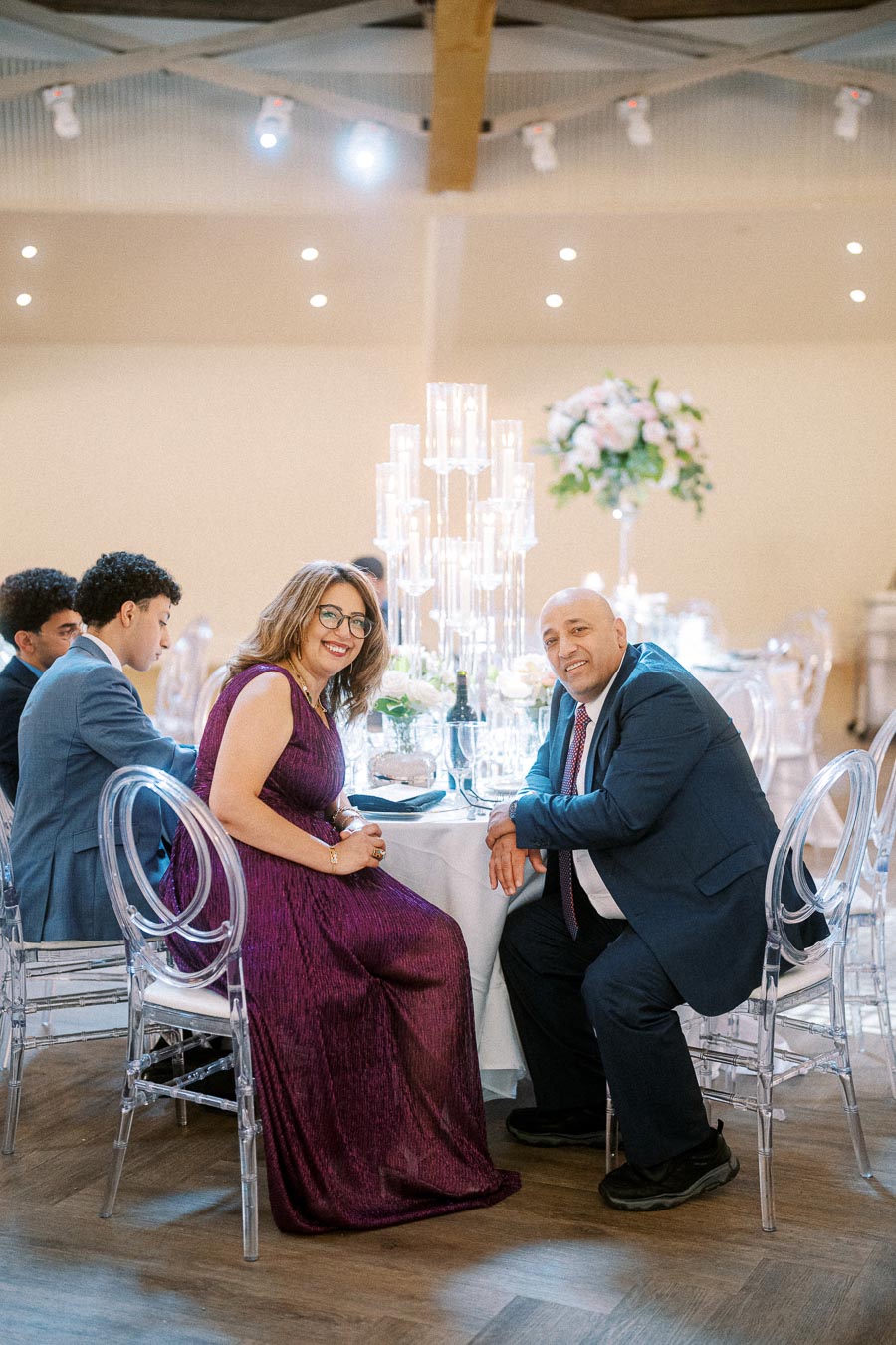 Elegant wedding reception with guests seated at a beautifully decorated table, featuring a tall candle centerpiece, floral