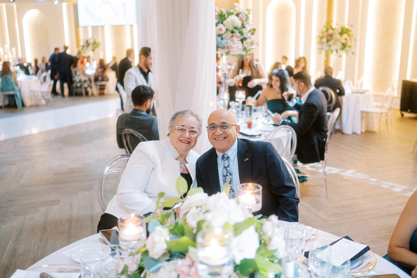 Elderly couple smiling at a decorated wedding reception table surrounded by guests, elegant lighting, and floral