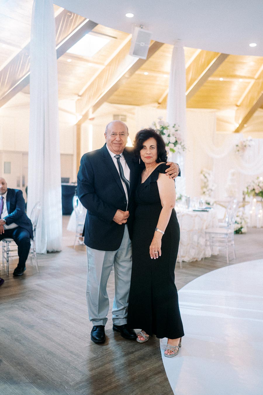 An elegantly dressed couple smiling and posing together in a beautifully decorated venue, featuring white drapery, floral