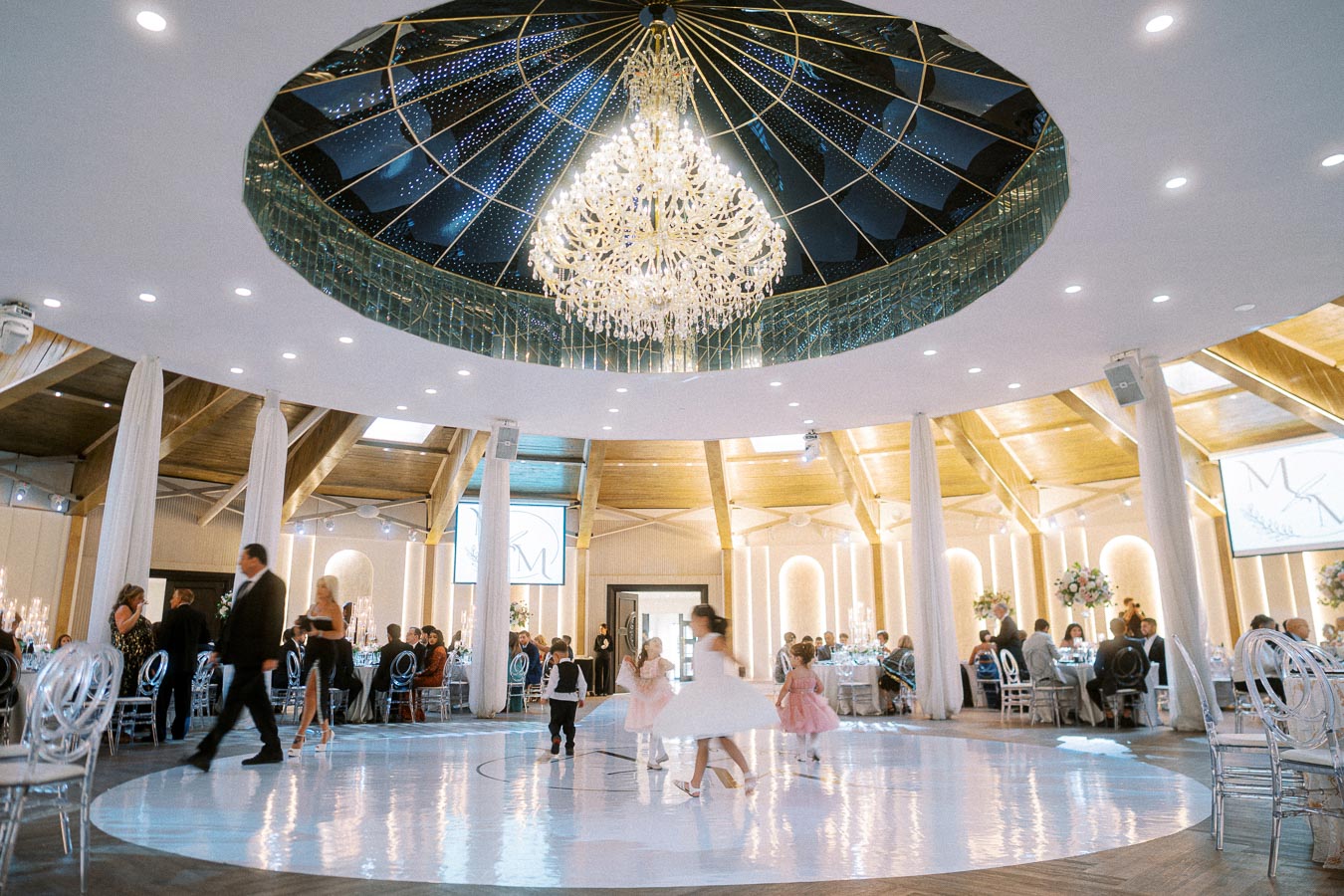 Elegant wedding reception with guests seated at round tables under a dazzling chandelier and children dancing on a white