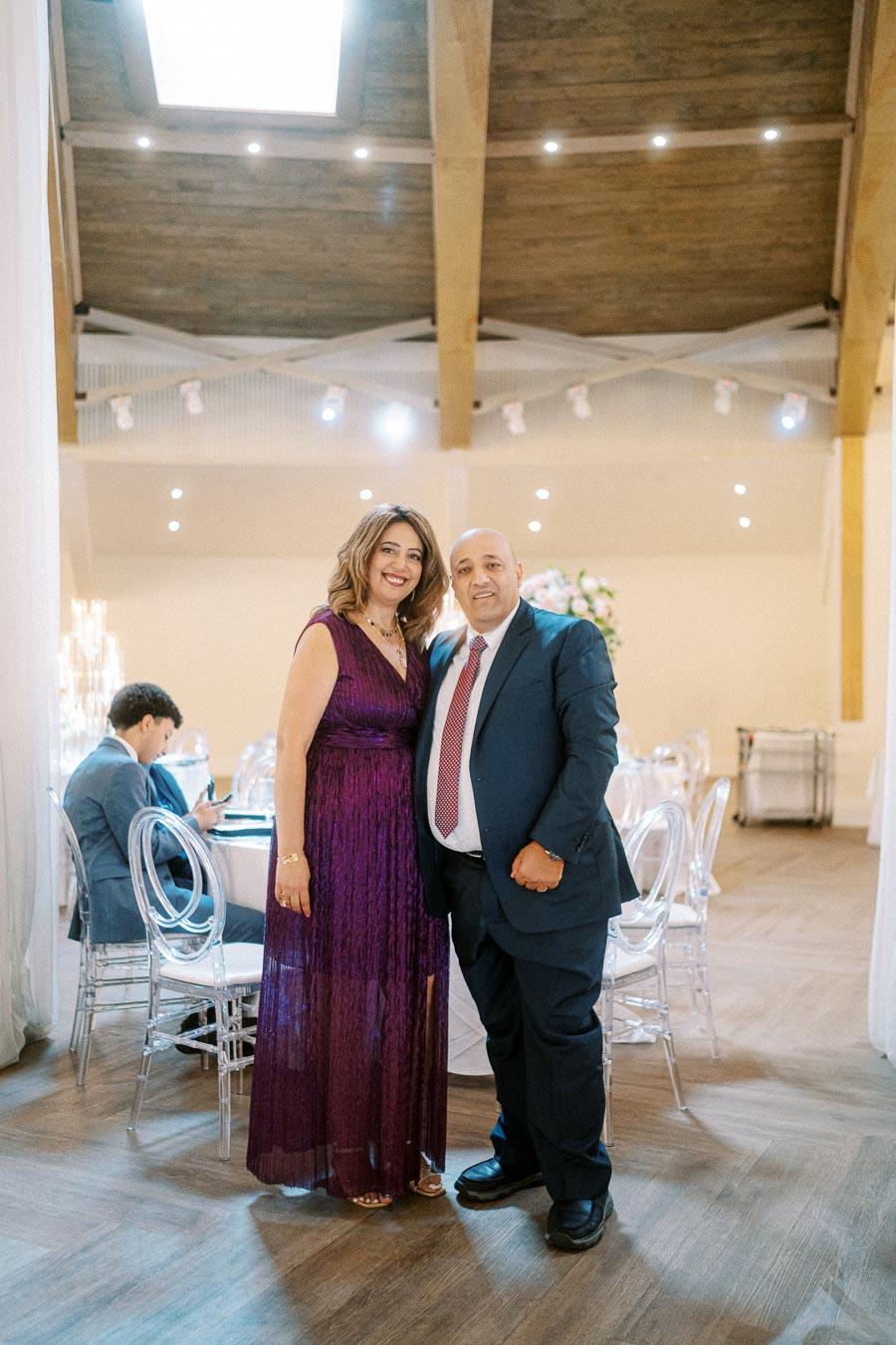 A smiling couple posing elegantly in a chic event hall, with the woman in a purple evening gown and the man in a suit and