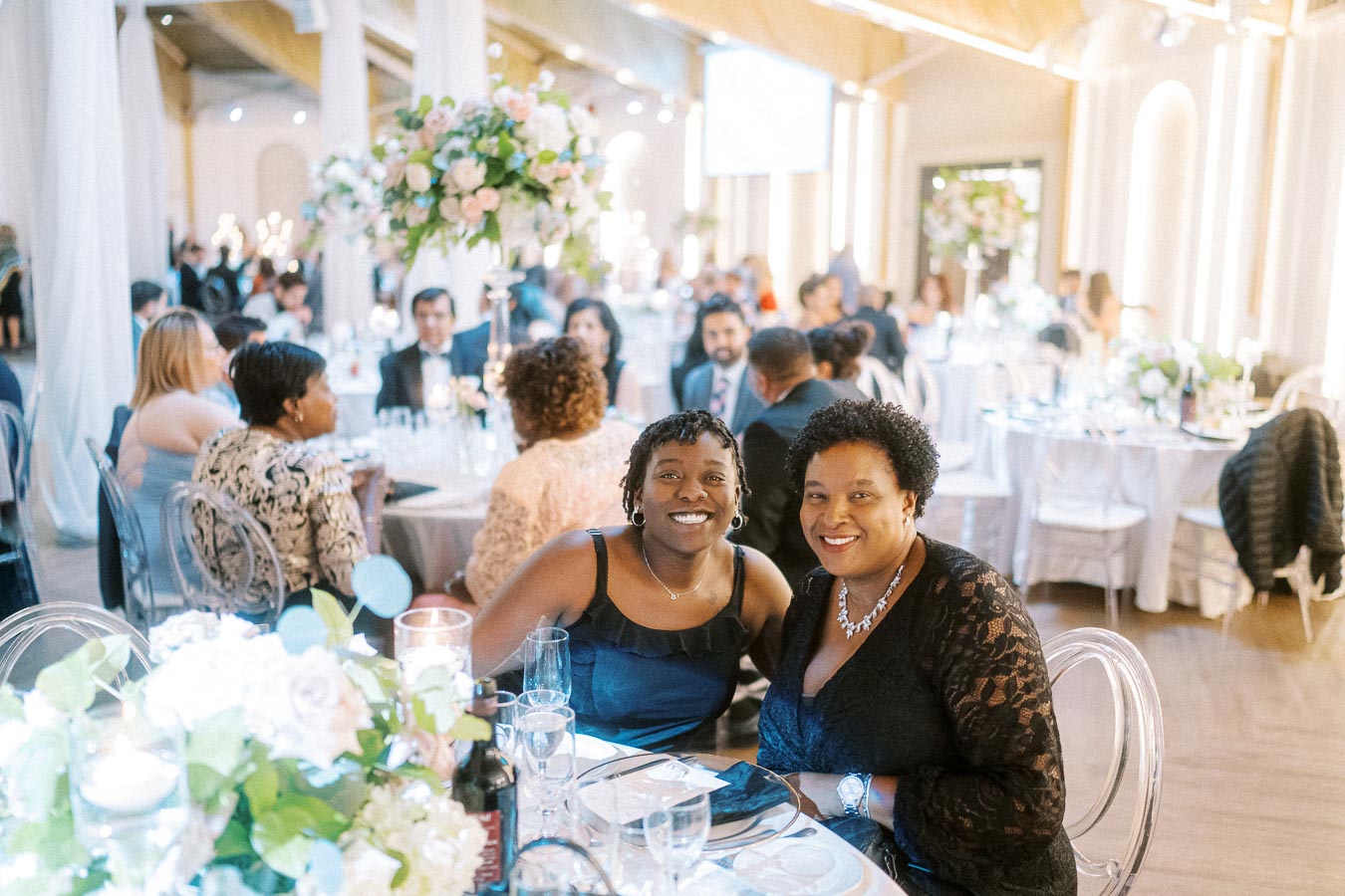 Two smiling women seated at a formal event surrounded by elegantly decorated tables with flowers and candles.