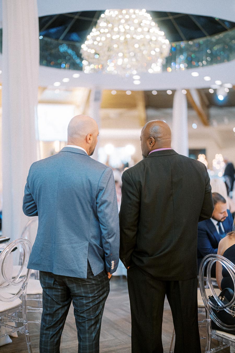 Two men in formal attire conversing under a large chandelier at an elegant event venue.