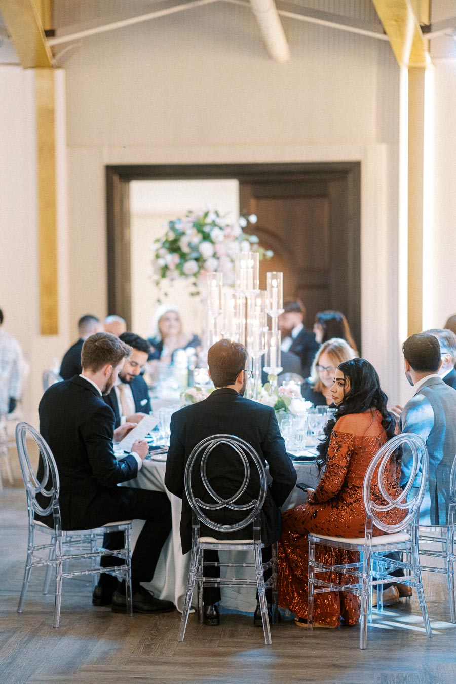 A group of elegantly dressed guests seated at a round table during a formal wedding reception, featuring clear chairs, a