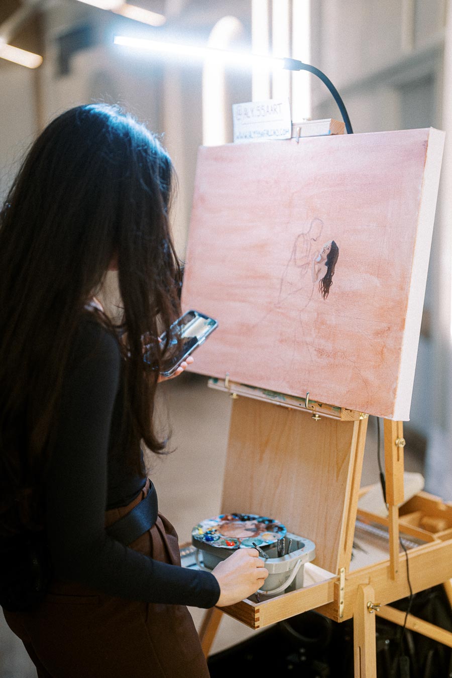 Female artist with long hair working on a canvas painting in a bright studio, using a smartphone for reference.