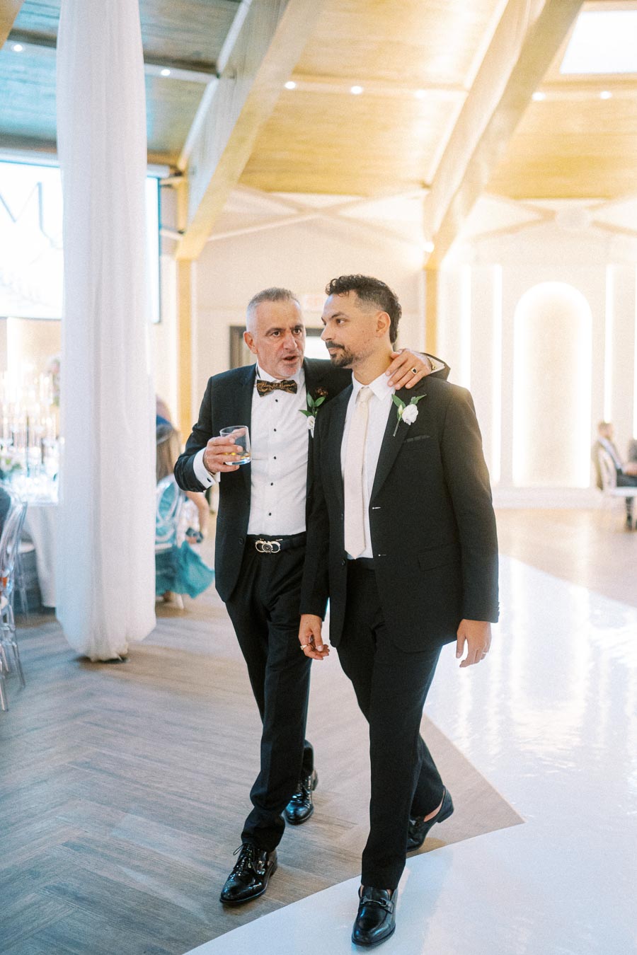 Two men in formal attire at a wedding reception. One has his arm around the other's shoulder, holding a drink, as they walk