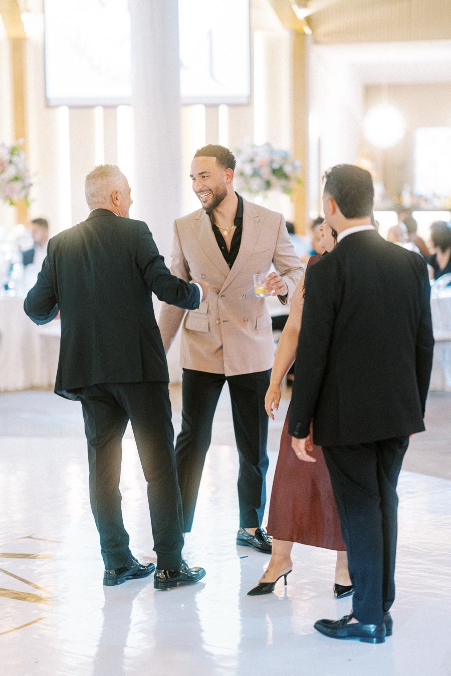 Four elegantly dressed individuals engage in conversation at a stylish indoor event, with a focus on a smiling man in a