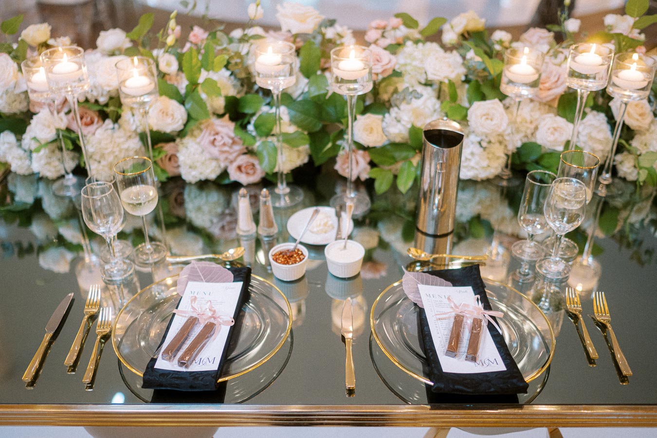 Elegant wedding table setting with gold cutlery, glassware, and floral centerpiece featuring white and blush flowers.