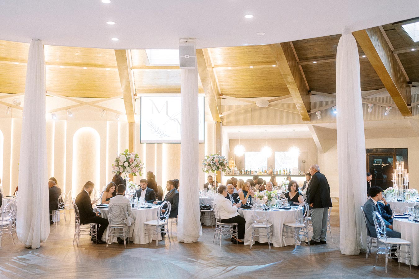 Elegant wedding reception in a modern, warmly lit venue with guests seated at round tables adorned with floral centerpieces.