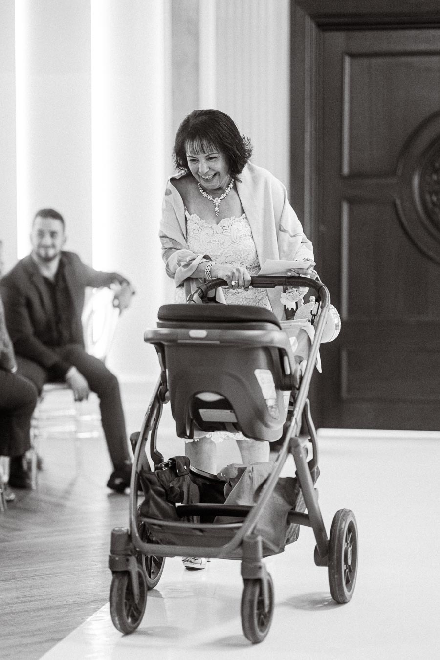 Black and white photo of a joyful woman pushing a stroller at an indoor event, with seated attendees in the background.