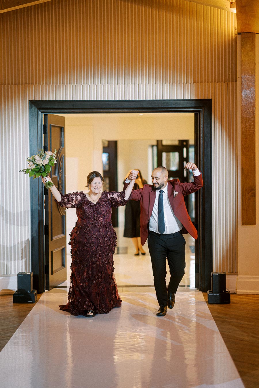 A joyful couple entering a wedding reception, both smiling and raising their hands in celebration. The woman is wearing a