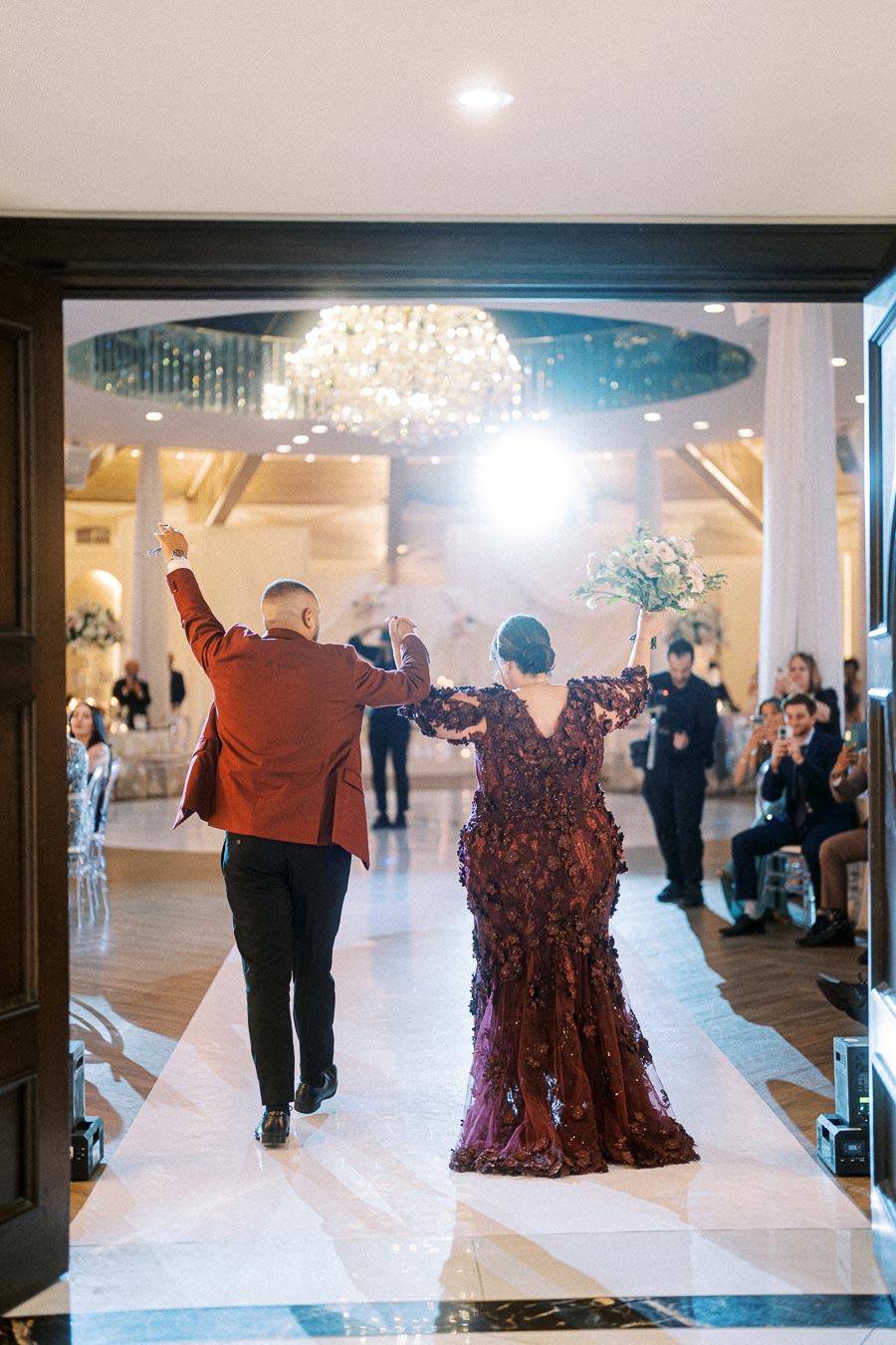 Couple joyfully enters elegant wedding reception venue, man in maroon suit and woman in ornate burgundy gown, greeted by