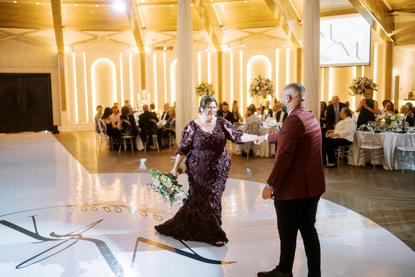 Elegant wedding reception dance floor, featuring a woman in a purple gown holding a floral bouquet, dancing with a man in a