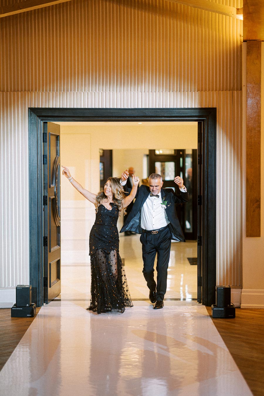 A couple joyfully enters a venue, dressed elegantly, with the woman in a black evening gown and the man in a suit,