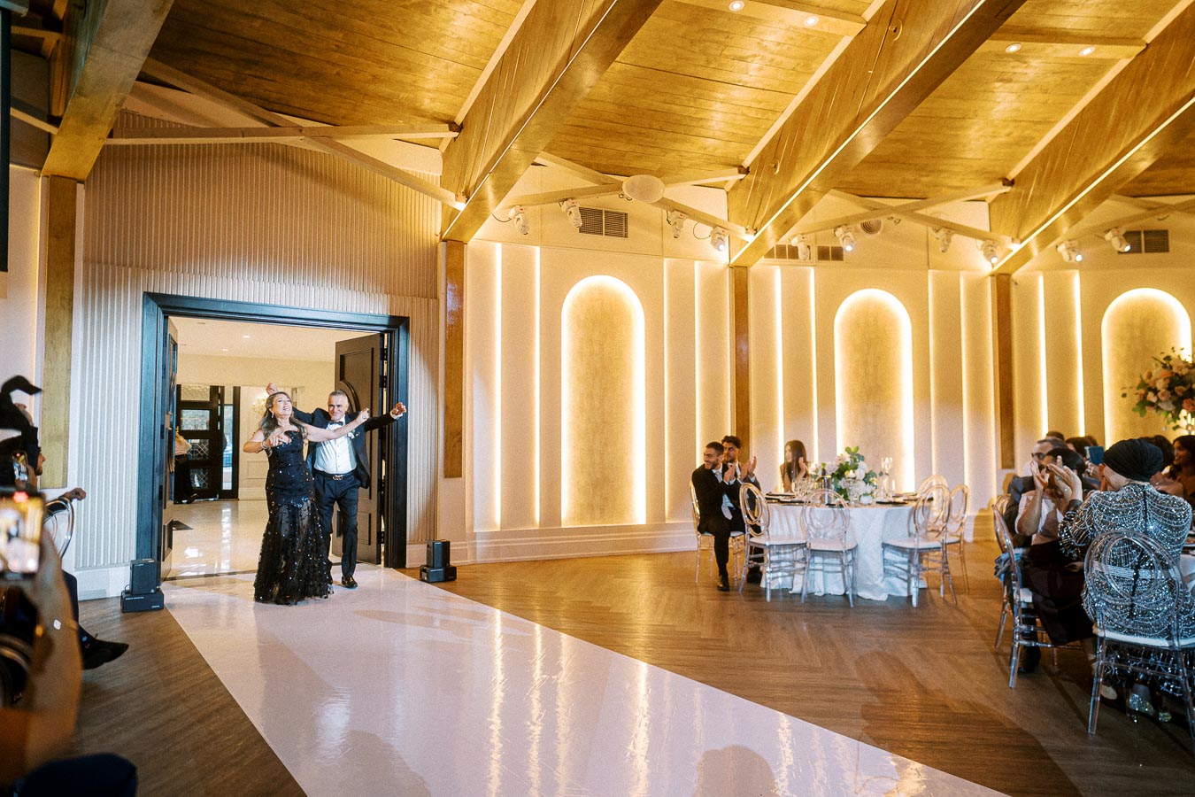 A couple enthusiastically enters a warmly lit, elegantly decorated wedding reception venue, with guests clapping and